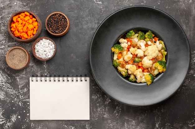 Top view broccoli and cauliflower salad in black bowl different spices and cutting carrot in bowls a notebook on dark surface