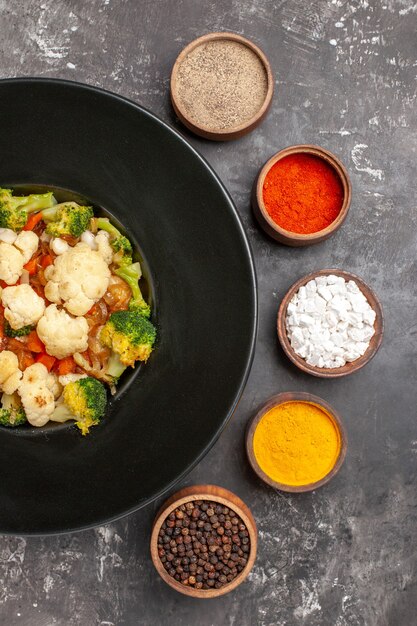 Top view broccoli and cauliflower salad on black oval plate different spices in small bowls on dark surface