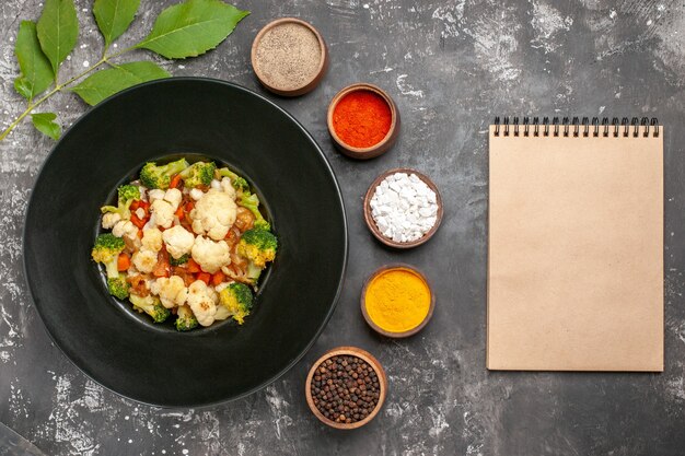 Top view broccoli and cauliflower salad on black oval plate different spices in small bowls a notebook on dark surface