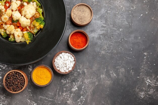 Top view broccoli and cauliflower salad on black oval plate different spices in small bowls turmeric sea salt black pepper on dark surface free place