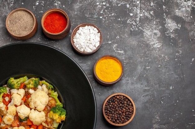 Top view broccoli and cauliflower salad on black oval plate different spices in small bowls turmeric sea salt peppers on dark surface free place