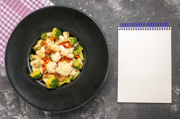 Top view broccoli and cauliflower salad on black oval plate pink and white checkered tablecloth a notepad on dark surface