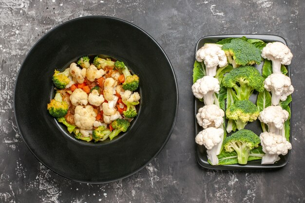 Top view broccoli and cauliflower salad on black oval plate raw broccoli and cauliflower on black rectangular plate on dark surface