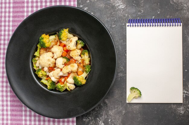 Top view broccoli and cauliflower salad on black oval plate on serving tray pink and white checkered tablecloth a notebook on dark surface