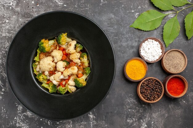 Top view broccoli and cauliflower salad on black oval plate on serving tray spices in small bowls on dark surface