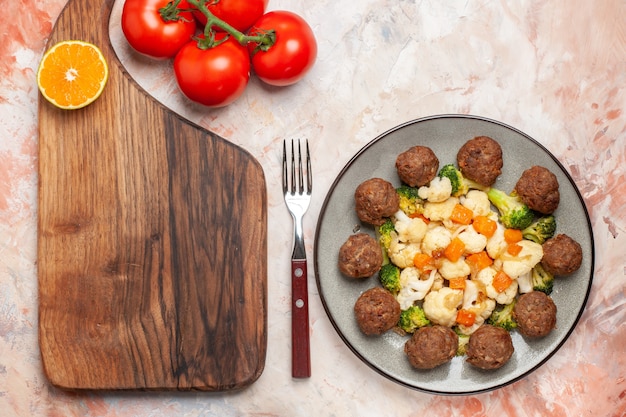 Top view broccoli and cauliflower salad and meatball on plate lemon slice on cutting board a fork tomatoes on nude background