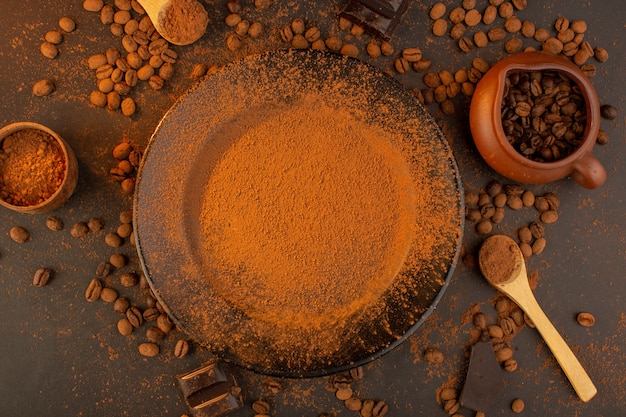 Una vista dall'alto semi di caffè marroni insieme a un piatto nero pieno di polvere di caffè con barrette di cioccolato dappertutto sullo sfondo marrone