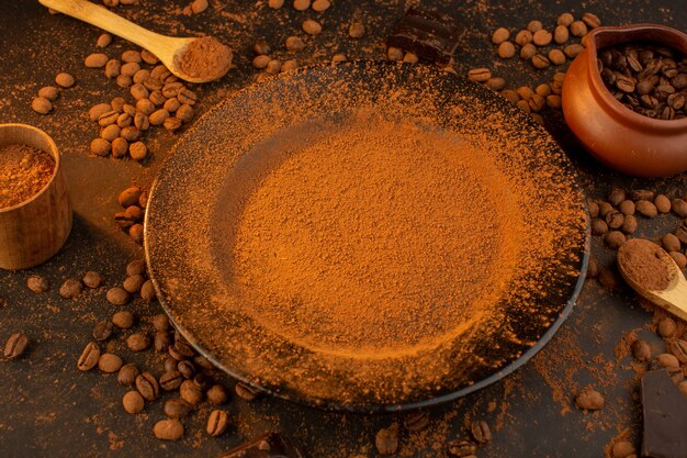 Una vista dall'alto di semi di caffè marroni insieme a un piatto nero pieno di polvere di caffè con barrette di cioccolato su tutto il tavolo marrone