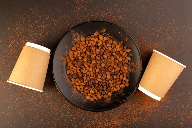 A top view brown coffee seeds inside black plate with empty cups on the brown table