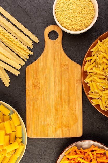 A top view brown desk wooden along with different formed yellow raw pasta inside bowls and plates isolated on the dark