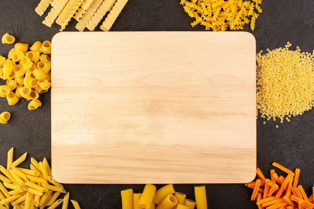 A top view brown desk wooden along with different formed yellow raw pasta isolated on the dark