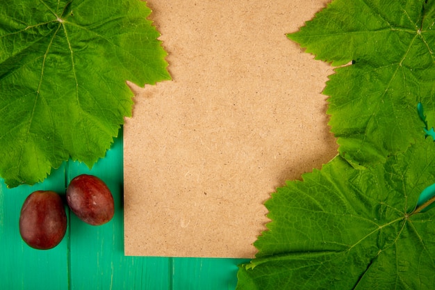 Top view of a brown paper sheet with sweet grapes and green grape leaves on green wooden table