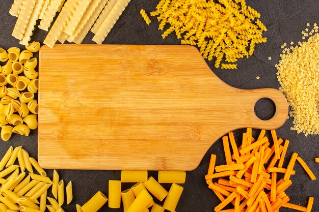 A top view brown wooden desk along with different formed yellow raw pasta isolated on the dark