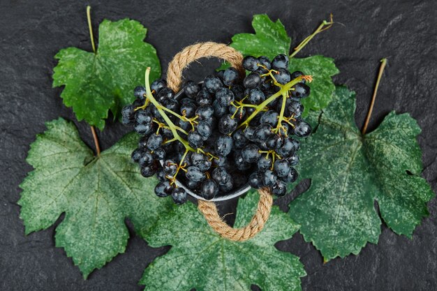 Top view of a bucket of black grapes with leaves on black surface