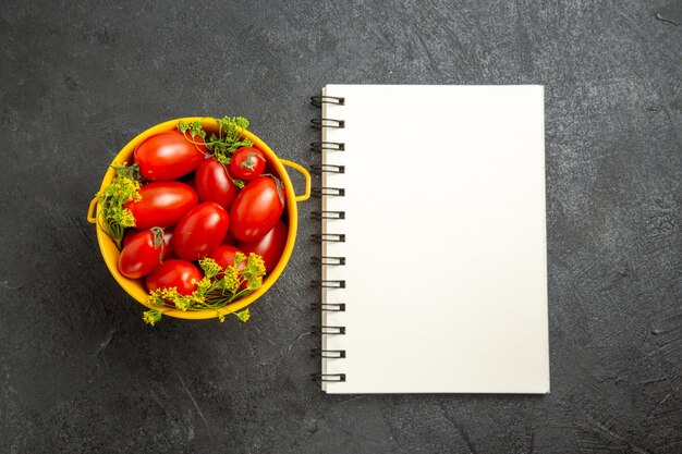 Top view bucket of cherry tomatoes and dill flowers and a notebook on dark background