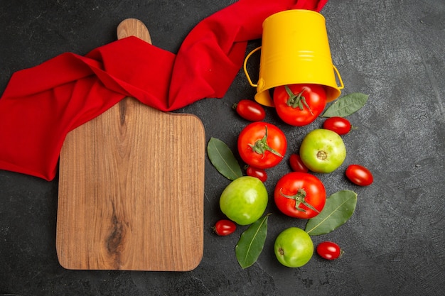 Top view bucket with red green and cherry tomatoes bay leaves red towel and a chopping board on dark background
