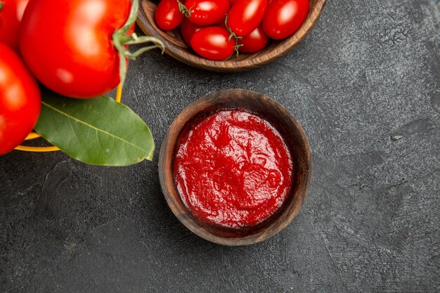 Top view a bucket with tomatoes and bay leaves bowls with cherry tomatoes and ketchup on dark ground