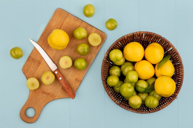 Top view of a bucket of yellow peaches with slices of green cherry plums on a wooden kitchen board with knife on blue background