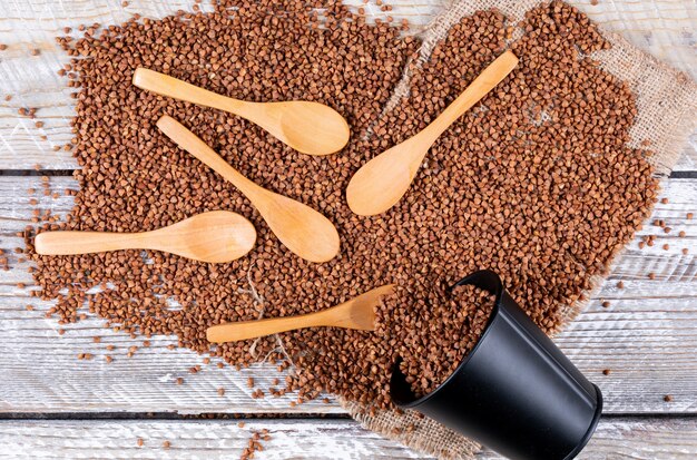 Top view buckwheats coming out of bucket with spoons on it on light wooden background. horizontal