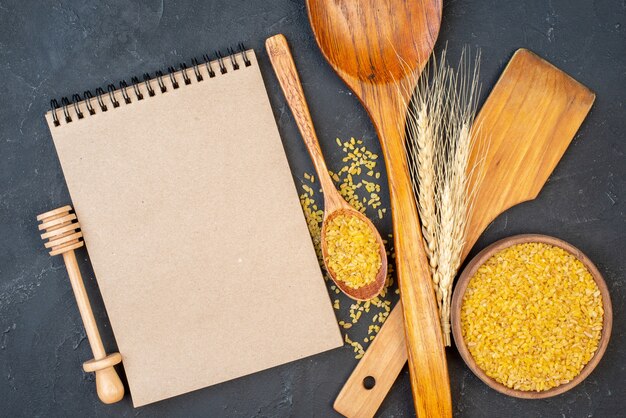 Top view bulgur wheat in bowl and wooden spoon two big wooden spoons honey stick notebook wheat spikes on table