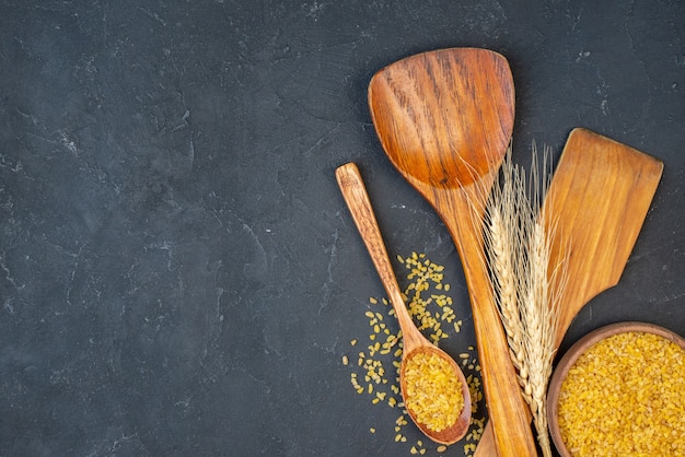 Free photo top view bulgur wheat in bowl and wooden spoon two big wooden spoons wheat spikes on black table with free space