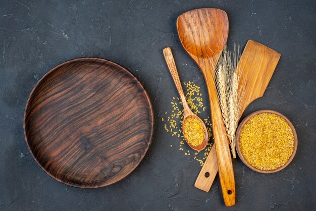 Top view bulgur wheat in bowl and wooden spoon two big wooden spoons wheat spikes wooden platter on table