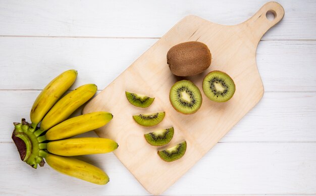 Top view of bunch of banana with slices of kiwi fruit on wooden cutting board on white rustic