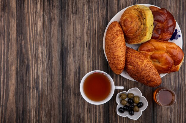 Вид сверху булочки на тарелке с чашкой чая с оливками на миске и медом на деревянном фоне с копией пространства