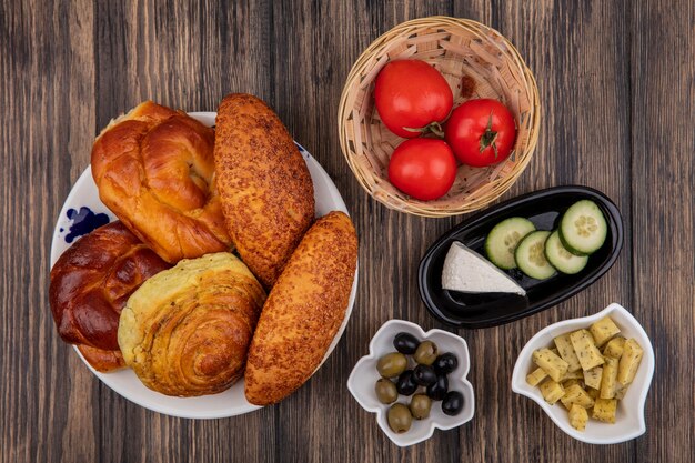 Top view of buns on a plate with fresh tomatoes on a bucket with olives on a bowl on a wooden background