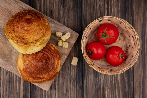 Top view of buns on a wooden kitchen board with chopped slices of cheese and tomatoes on a bucket on a wooden background