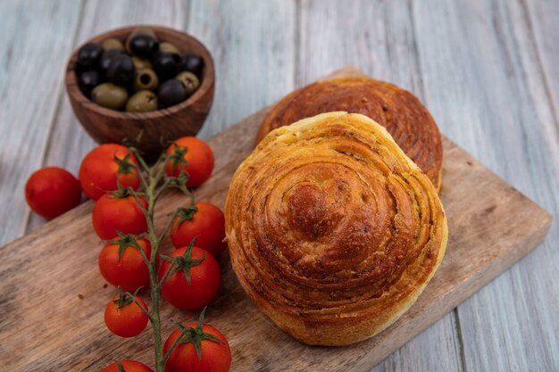 灰色の木製の背景の上の木製のボウルにオリーブとオリーブと新鮮なブドウのトマトと木製のキッチンボード上のパンの上面図
