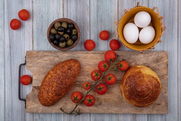 Top view of buns on a wooden kitchen board with vine tomatoes with olives and eggs on a grey wooden background