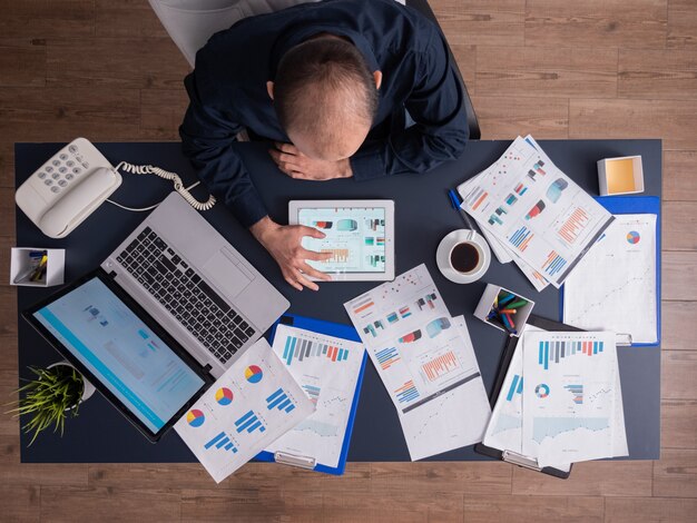 Top view of businessman using tablet pc analyzing financial charts and documents, sitting at desk in corporate office