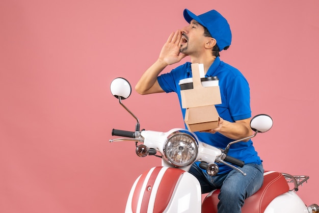 Foto gratuita vista dall'alto di un corriere impegnato che indossa un cappello seduto su uno scooter in possesso di ordini che chiama qualcuno su sfondo color pesca pastello