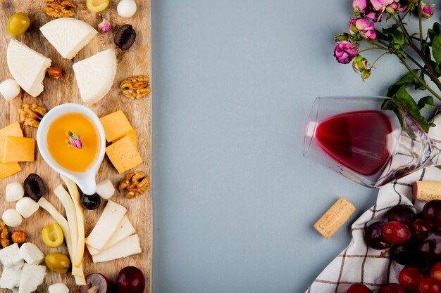 Top view of butter with cheese grape olive nuts on cutting board and glass of wine corks flowers on white with copy space