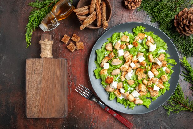 Top view caesar salad on oval plate fork bowl with dried crust chopping board on dark red background