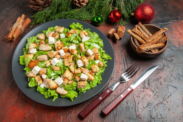 Top view caesar salad on oval plate fork knife bowl with dried crust xmas tree toys cinnamon sticks on dark red background