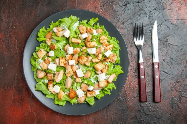 Top view caesar salad on oval plate fork and knife on dark red background