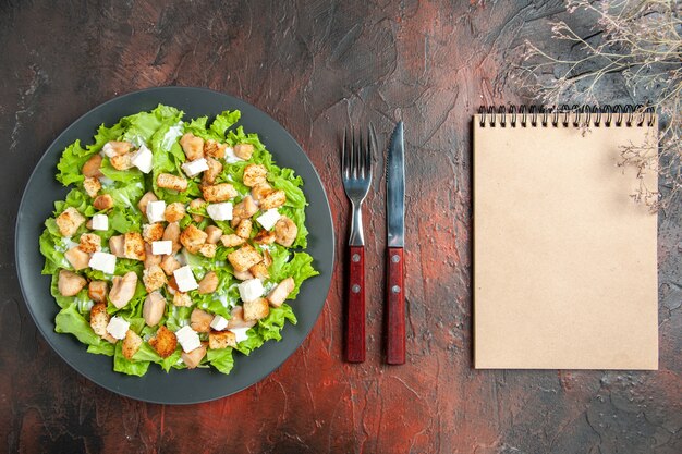 Вид сверху салат цезарь на овальной тарелке, вилка и блокнот с ножом на темно-красном фоне