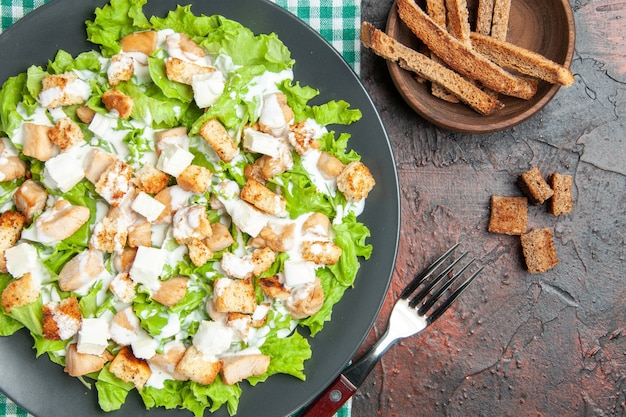 Top view caesar salad on oval plate green white checkered napkin fork on dark red background