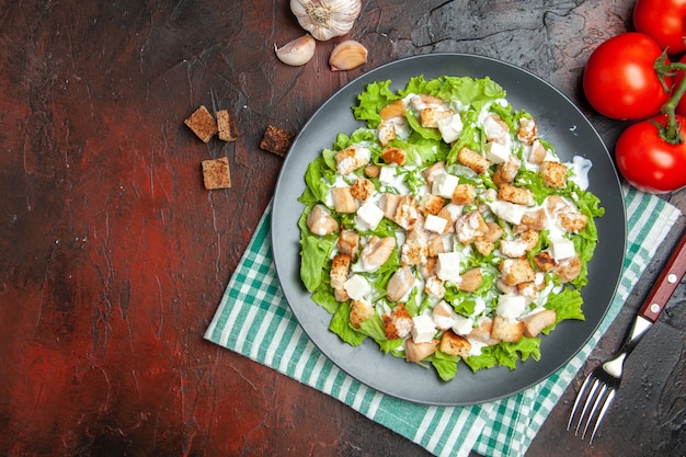 Top view caesar salad on oval plate green white checkered napkin tomatoes fork on dark red background