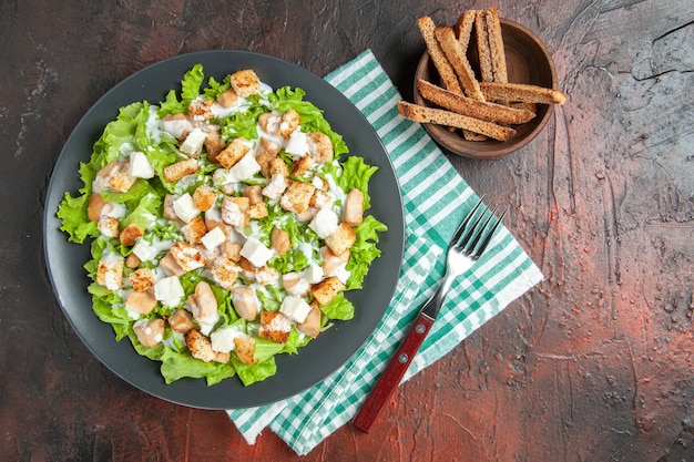 Top view caesar salad on oval plate green white checkered tablecloth bowl with dried crust fork on dark red background