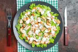 Free photo top view caesar salad on oval plate green white checkered tablecloth fork and knife on dark red background