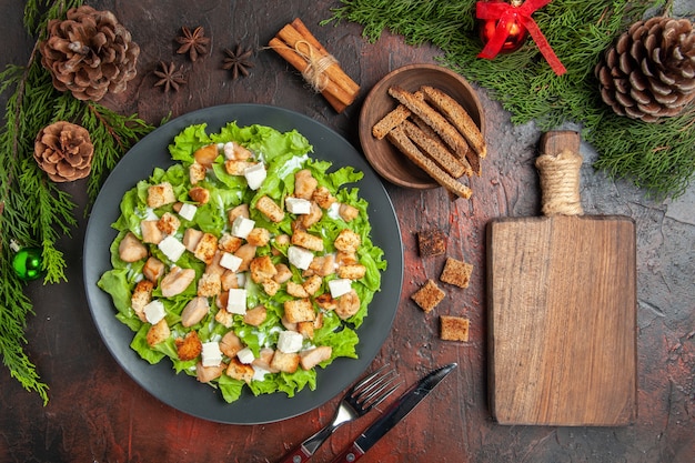 Top view caesar salad on oval plate pine tree branches fork knife anises bowl with dried crust chopping board on dark red background