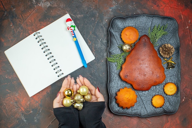 女性の手のノートの黒いプレートのクリスマスボールのおもちゃの上面図のケーキとカップケーキと濃い赤のテーブルのペン 無料写真