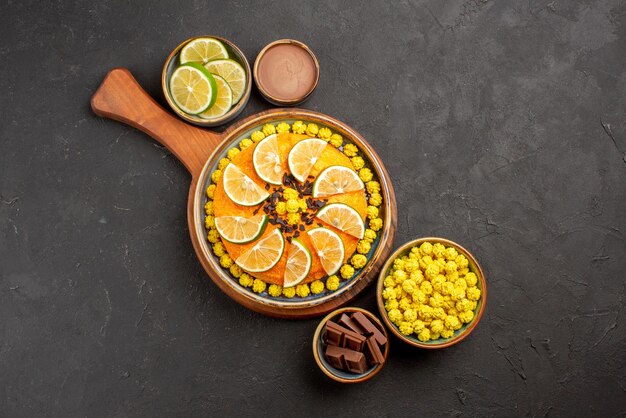 Top view cake on board candies chocolate cream and sliced limes in bowls and orange cake on the cutting board on the table