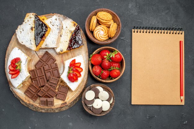 Top view cake slices with fruits cookies and choco bars on dark surface