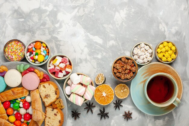 Top view of cake slices with macarons, bagels and candies with cup of tea on white table