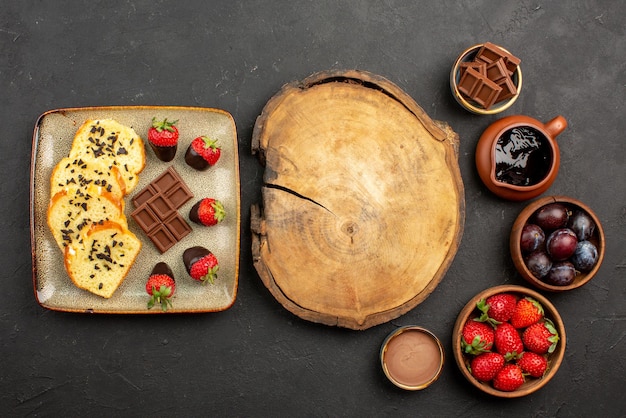 Top view cake and strawberries cutting board between pieces of cake with chocolate on the left and bowls with strawberries berries and chocolate sauce on the right side of the table