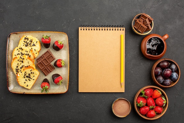 Top view cake and strawberries notebook and pencil between pieces of cake with chocolate on the left and bowls with strawberries berries and chocolate sauce on the right side of the table